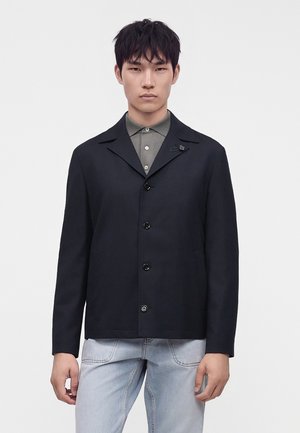Junger Mann mit kurzen schwarzen Haaren, der eine schwarze, geknöpfte Jacke über einem grauen Hemd mit Kragen und hellblauen Jeans vor einem schlichten weißen Hintergrund trägt.