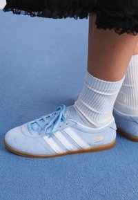 Gaiši zili zamšaini Adidas Gazelle sporta apavi ar baltām svītrām un auklām; kopā ar ribotām baltām zeķēm. Fons ir zils.