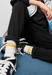 Pantaloni jogger neri, sneakers neri con accenti bianchi e calzini bianchi con strisce gialle. Texture liscia, vestibilità comoda, look casual.