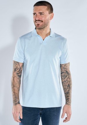 Mann mit kurzen Haaren und Tätowierungen auf beiden Armen, der ein hellblaues Poloshirt und dunkle Jeans trägt und zur Seite schaut.