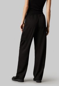 Sorte brede bukser med snøre i taljen og sidelomme, der har en glat tekstur og lige snit, set fra ryggen.