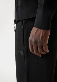 Zwarte sportjack met een gestructureerde afwerking, voorzien van een ritssluiting aan de voorkant en een trekkoord. Zwarte broek heeft een driehoekig merklogo aan de zoom.