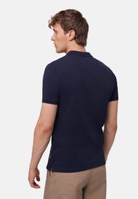 Laivastonsininen lyhythihainen polo-paita, puuvillamateriaalia, litteä kaulus, sivuhalkiot ja pieni punainen yksityiskohta helmassa.