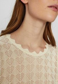 Pletený svetr s vroubkovaným výstřihem, v krémové barvě, se vzorem srdce vytvořeným pomocí otvorových detailů po celé látce.