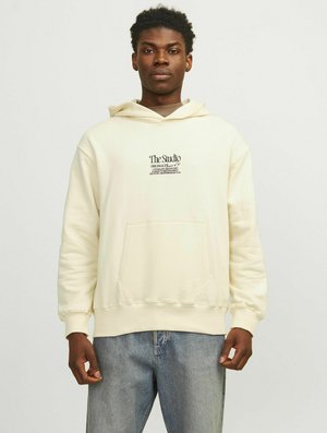 Cremfarbener Hoodie aus weichem Stoff, mit einer Fronttasche und einem grafischen Druck auf der Brust. Kombiniert mit hellblauen Jeans.