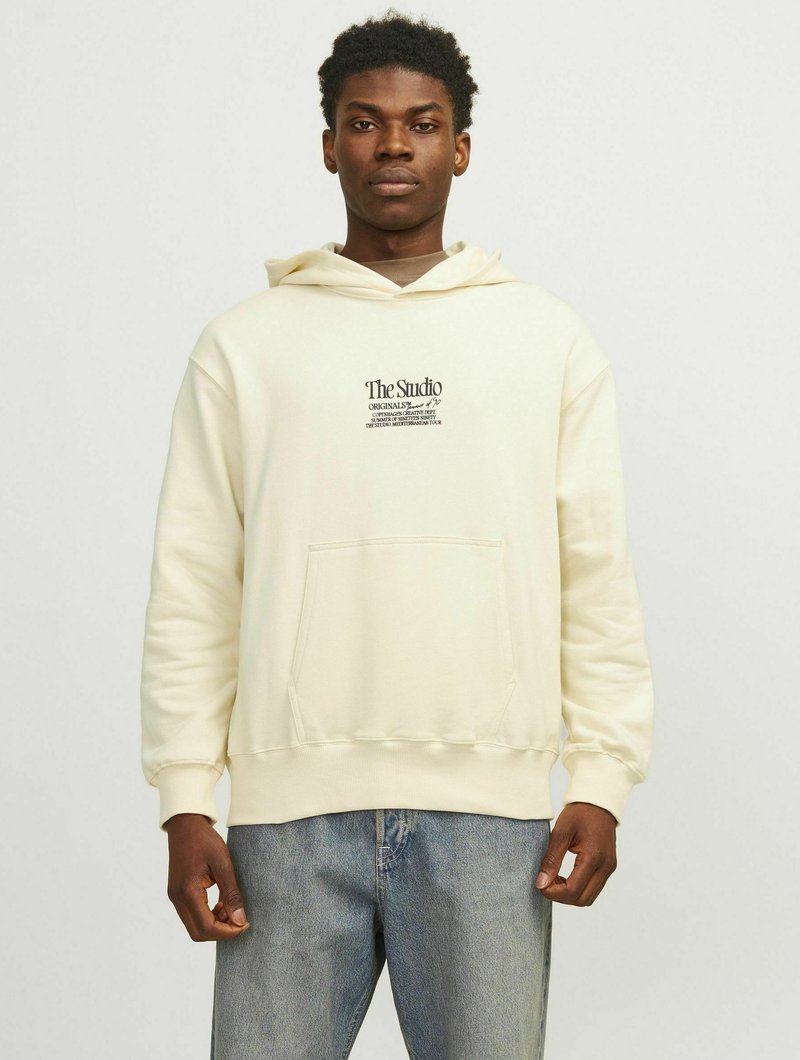 Sweat à capuche couleur crème en tissu doux, avec une poche avant et un imprimé graphique sur la poitrine. Associé à un jean bleu clair.