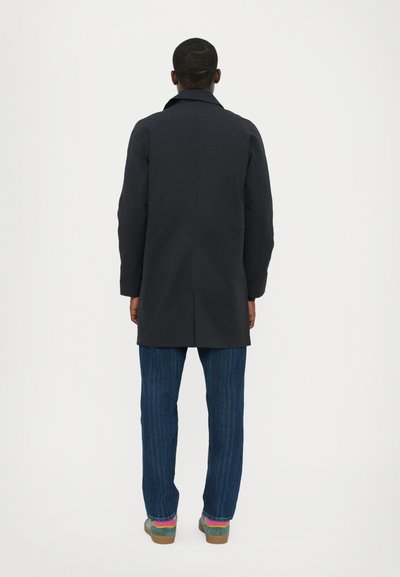 Zwarte, knielange jas met een rechte silhouet, op maat gemaakte kraag en zichtbare naden, gecombineerd met gestreepte blauwe jeans en contrasterende sokken.