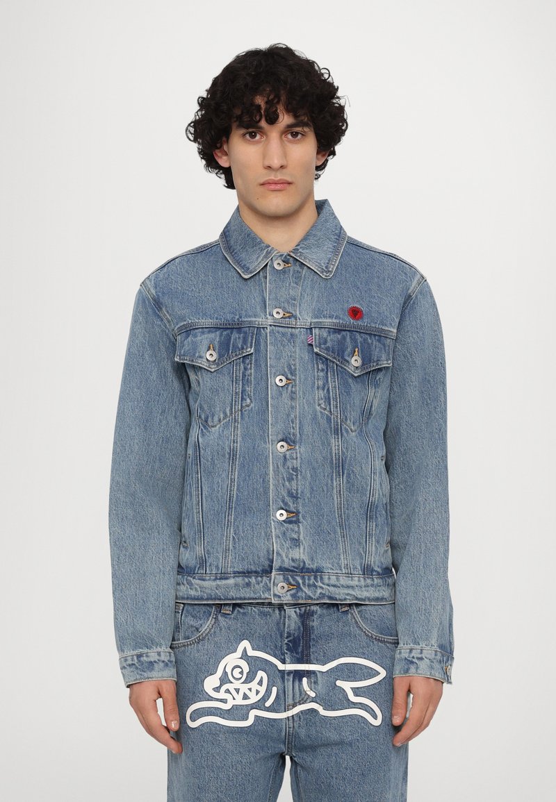 Giacca di jeans di colore azzurro chiaro con colletto a punta, due tasche sul petto, bottoni argentati e un emblema rosso sul lato sinistro. Pantaloni in denim abbinati in basso.