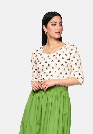 Frau mit dunklem Haar, die ein cremefarbenes Oberteil mit kleinen Blumenmustern und einen hellgrünen Rock mit hoher Taille trägt, blickt nach links.