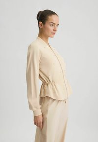 Veste beige zippée à manches longues, taille froncée et embellissements décoratifs. Texture lisse et coupe décontractée.