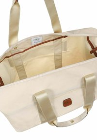 Borsa tote in tela beige con manici in nastro dorato, chiusura con zip e accenti in pelle marrone. L'interno presenta una fodera chiara e nessuna tasca.