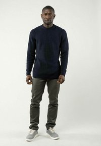 Marineblauer, gerippter Strickpullover, taillierte graue Hose und graue Sneakers mit cremefarbenen Schnürsenkeln. Steht vor einem einfarbigen Hintergrund.