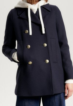 Femme portant un manteau croisé bleu marine avec des boutons métalliques sur un sweat à capuche blanc et un jean bleu, une main dans la poche.