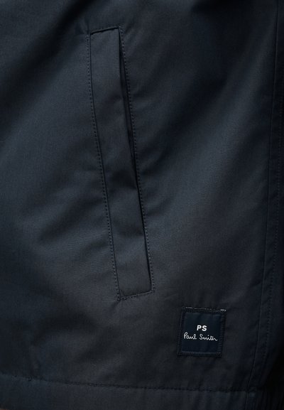 Κοντινή λήψη μαύρου υφασμάτινου σακακιού με κάθετη τσέπη σχισμή και μικρή τετράγωνη ετικέτα με την ένδειξη "PS Paul Smith" κοντά στο κάτω τελείωμα.