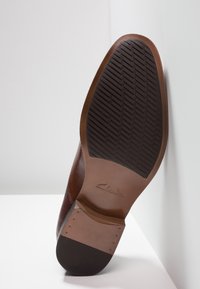 Chaussure en cuir marron avec une tige texturée et une semelle en caoutchouc. La semelle présente un motif sur la bande de roulement et un logo embossé sur le côté.