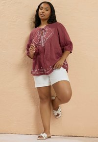 Roestkleurige, losvallende blouse met witte borduursels en strikdetail, gecombineerd met witte shorts en witte instapsandalen.