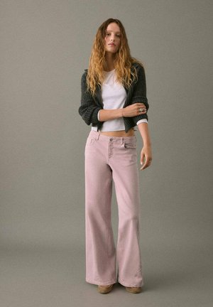Pantalones de mezclilla acampanados de color rosa claro con una cintura alta, cinco bolsillos y costuras decorativas. Combinados con un cárdigan gris corto y una blusa blanca.