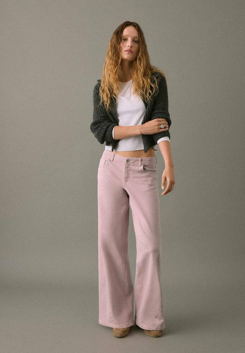 Pantalones de mezclilla acampanados de color rosa claro con una cintura alta, cinco bolsillos y costuras decorativas. Combinados con un cárdigan gris corto y una blusa blanca.