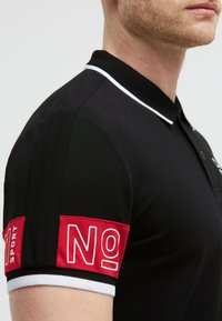 Polo nero con colletto bianco, dotato di dettagli rossi sulle maniche con il testo "NO" e "SPORT". Tessuto strutturato, design aderente.