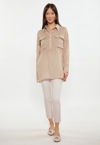 Camicia tunica di lino beige con patta dei bottoni, due tasche sul petto e spacchi laterali, abbinata a pantaloni slim chiari e scarpe basse.