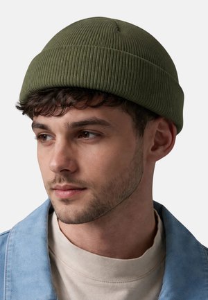 Junger Mann mit leichtem Gesichtshaar, der eine olivgrüne, gerippte Strickmütze und eine hellblaue Jeansjacke über einem beigen Rundhalsausschnitt trägt.