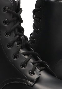 Botas de combate de cuero negro con ojales circulares, cordones negros y una textura suave. Presentan un diseño de caña alta y suela resistente.