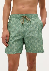 Shorts de bain verts à motifs, avec une coupe décontractée, une taille élastique et un cordon de serrage marron. Le design présente des motifs floraux et des lignes ondulées.