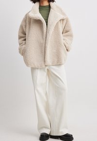 Persoon in een beige fluffy jas over een olijfgroene top, met crèmekleurige wijde broek en zwarte chunky schoenen, staande met handen in de zakken.