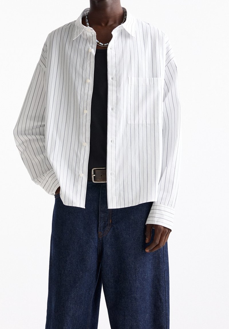 Personne portant une chemise blanche à manches longues avec des rayures verticales noires sur un haut noir, un jean bleu foncé et un collier de perles.