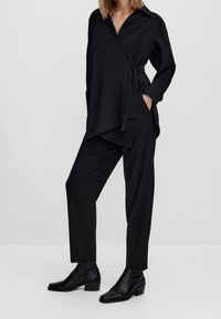 Musta kietaisupaita, jossa on kauluskäänne ja sivusolmu, yhdistettynä vastaaviin kapeisiin housuihin ja nilkkureihin, esittelee tyylikkään siluetin.