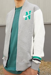 Giacca grigia con zip e maniche bianche, caratterizzata da un logo "N" ricamato verde e lo swoosh di Nike. Include polsini a costine con strisce colorate.