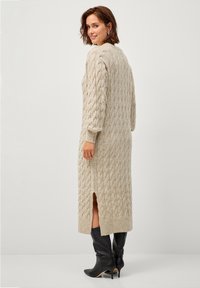 Beiges Strickkleid aus Kabelmuster mit langen Ärmeln, seitlichen Schlitzen und gerippten Bündchen. Kombiniert mit kniehohen schwarzen Stiefeln mit Absatz.