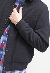 Schwarze Bomberjacke mit strukturierter Oberfläche, Reißverschluss und Druckknopfverschluss. Verfügt über gerippte Bündchen und Saum, getragen über einem karierten Hemd.