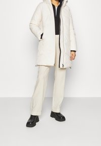 Cappotto bianco gonfio con cappuccio, chiusura con zipper e motivo a trapunta diagonale, abbinato a pantaloni crema e stivali con zeppa neri.