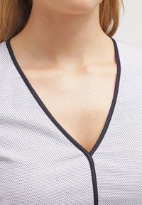 Šedý top s výstřihem do V a texturovaným síťovaným vzorem, s tmavým lemováním podél výstřihu a knoflíkovými detaily. Hladká tkanina jemně splývá.