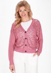 Rosa Cardigan mit Pailletten-Design, gerippten Kanten und V-Ausschnitt. Knopfleiste, lockere Passform und strukturierte Ärmel. Über einem weißen Oberteil getragen.