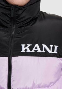 Polstret jakke med en sort topsektion og lyse lilla bund. Den har en høj krave, lynlås lukning og broderet "KANI" logo.