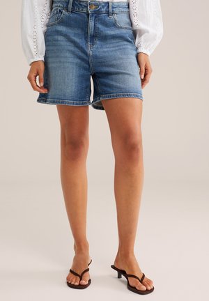 Džínové šortky nosené s bielou blúzkou s dlhými rukávmi a čiernymi sandálmi na nízkom opätku, zobrazujúce nohy od polovice stehien až po chodidlá.