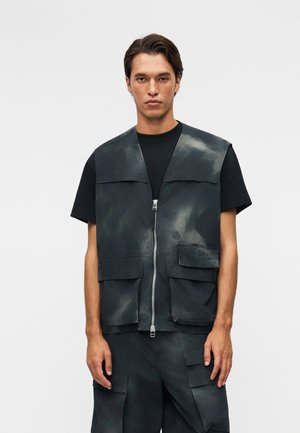 Νεαρός άνδρας που φοράει μαύρο κοντομάνικο μπλουζάκι με σκοτεινό γκρι γιλέκο εργασίας με φερμουάρ και αντίστοιχο cargo παντελόνι, στέκεται μπροστά σε απλό φόντο.