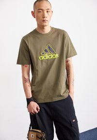 T-shirt en coton vert olive avec un grand logo Adidas texturé en jaune et bleu marine. Manches courtes et coupe décontractée.