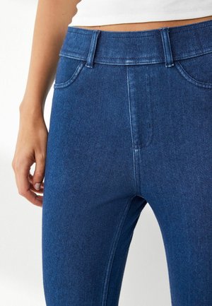 Tmavě modré džíny z denimu s středním pasem, pěti-kapsovým designem a hladkou strukturou. Tkanina vykazuje jemné detaily prošívání.