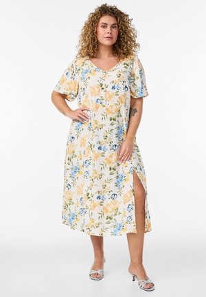 Kobieta o kręconych włosach w kremowej sukience midi z żółto-niebieskim kwiatowym nadrukiem i rozcięciem po boku, pozująca z ręką na biodrze w butach na obcasie z odkrytymi palcami.