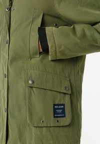 Oliņu zaļš žakets, izgatavots no teksturēta auduma ar spiedpogām, sānslēdzēju kabatu un brenda etiķeti priekšpusē. Ietver melnus aproces.