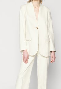 Femme portant un tailleur écru avec un blazer à un seul bouton doré et un pantalon large assorti, debout devant un fond uni.