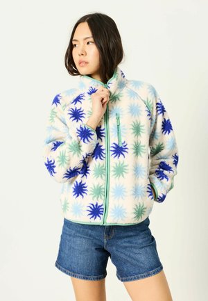 Frau trägt eine Fleecejacke mit Reißverschluss mit blauen und grünen Sternexplosionsmustern und Jeansshorts, schaut zur Seite und hält eine Hand am Kragen.