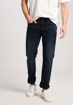 Mann trägt hochgekrempelte dunkelblaue Jeans, weiße Turnschuhe und ein weißes kurzärmeliges T-Shirt, steht vor einem einfarbigen Hintergrund.