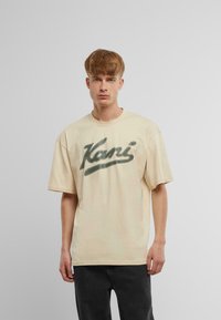 Beige kortærmet T-shirt med et stort grønt grafisk logo på forsiden. Stoffet virker blødt, og T-shirten har en afslappet, boxy pasform.