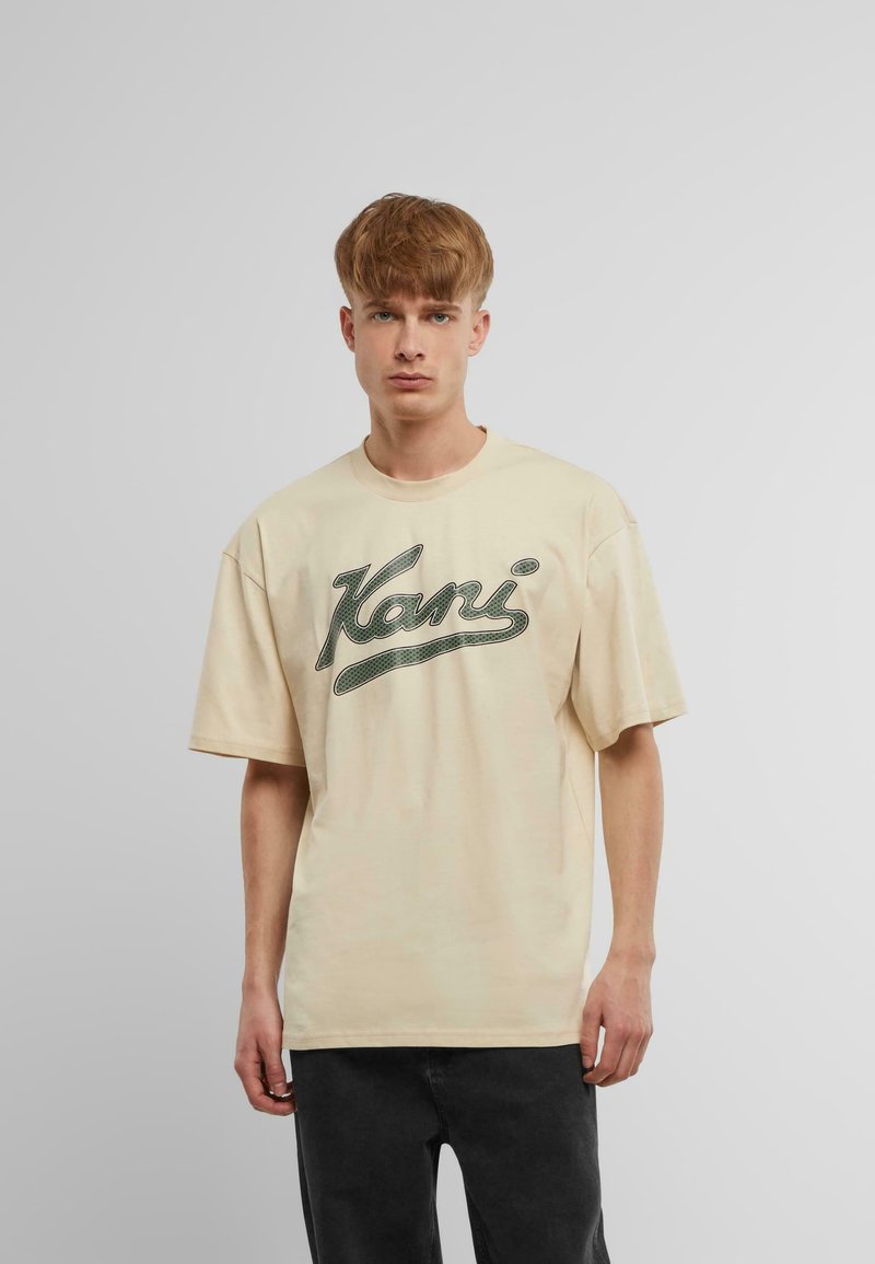 Beige kortærmet T-shirt med et stort grønt grafisk logo på forsiden. Stoffet virker blødt, og T-shirten har en afslappet, boxy pasform.