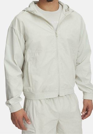 Mann trägt eine hellbeige Kapuzenjacke mit Reißverschluss, elastischen Bündchen und passende Hosen vor einem schlichten Hintergrund.