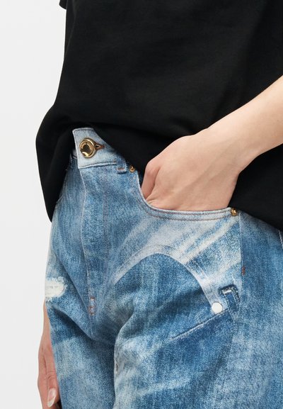 Χέρι που ξεκουράζεται μέσα στην μπροστινή τσέπη μπλε τζιν με όξινη πλύση, σε συνδυασμό με μαύρο πουκάμισο.